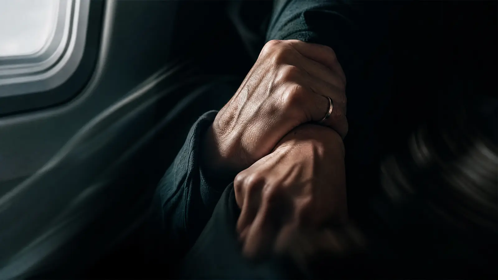A woman's hands gripping a man's forearm against the bright light of an aircraft window, knuckles white, refusing to let go.
