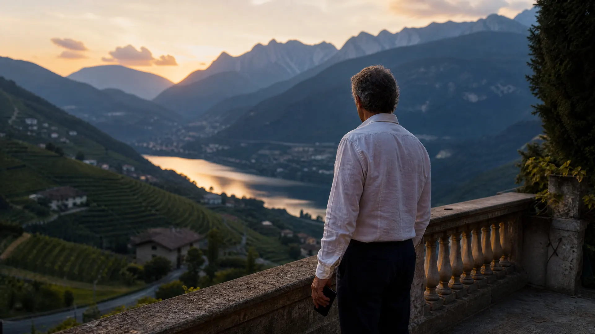 A man stands alone on an alpine terrace at dusk, looking out over an Italian valley as the light fades.